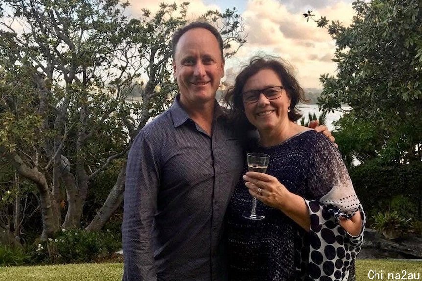 Tim stands with his arm around Lynelle in a garden, she holds a glass of wine. Water can be seen in the distance behind them.