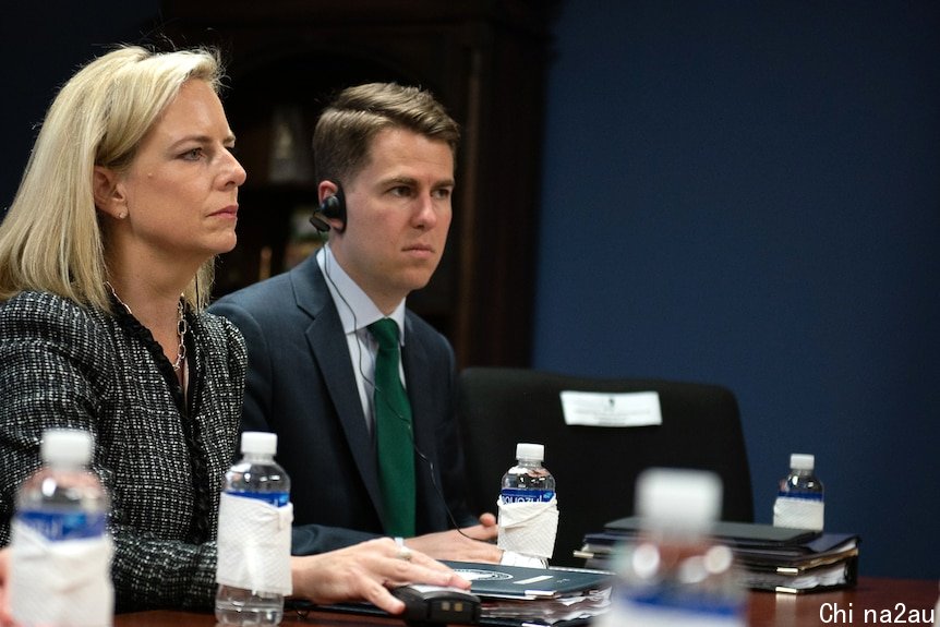 Miles Taylor sits next to a woman at a table. He has an earpiece on.