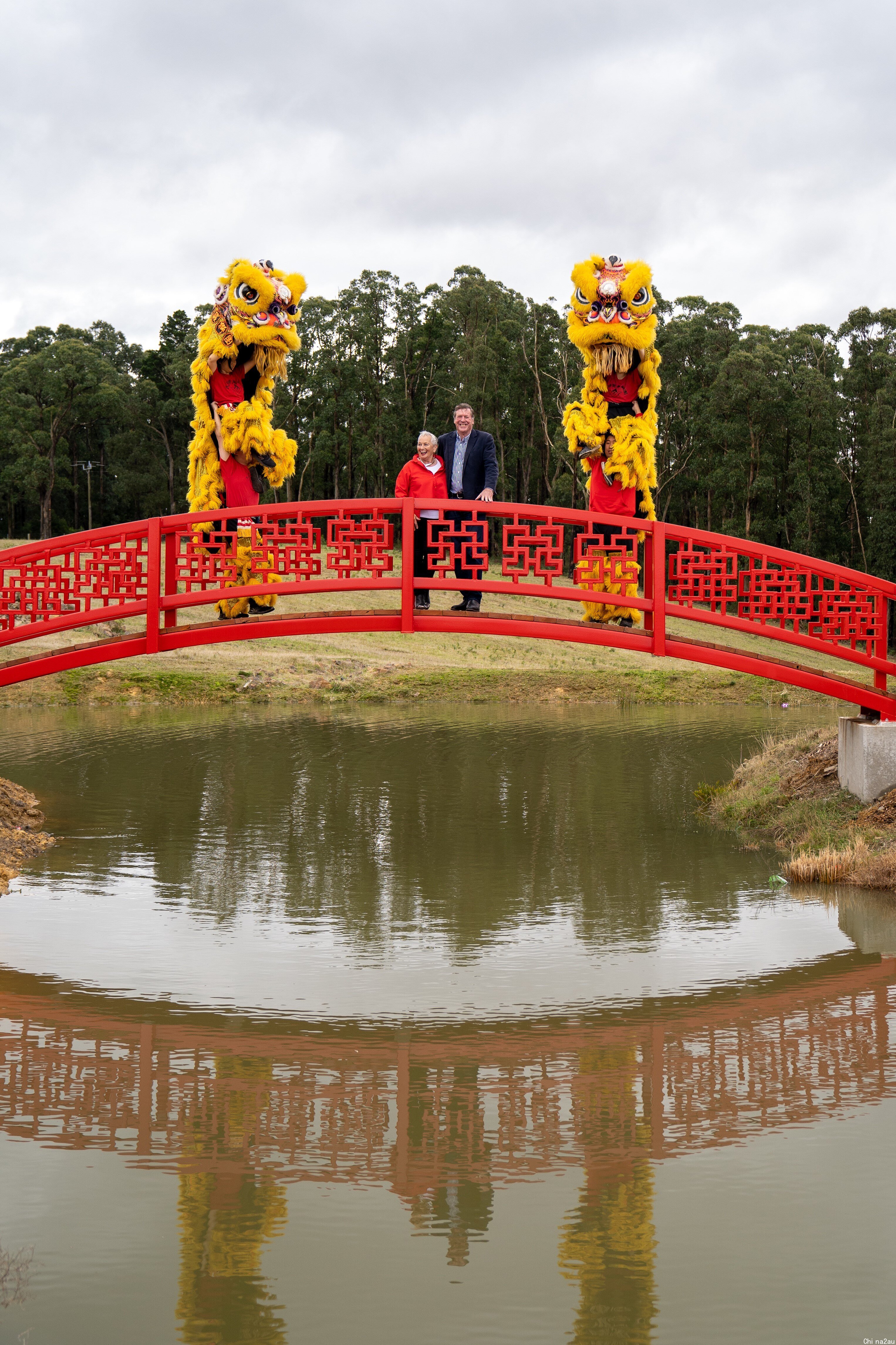 一堆夫妇站在中式拱桥上，两侧有舞狮队。