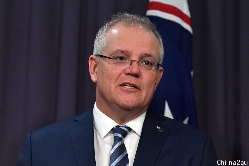 Scott Morrison looks to the right as he stands in front of a blue background mid-speech. An Australian flag is also behind him.