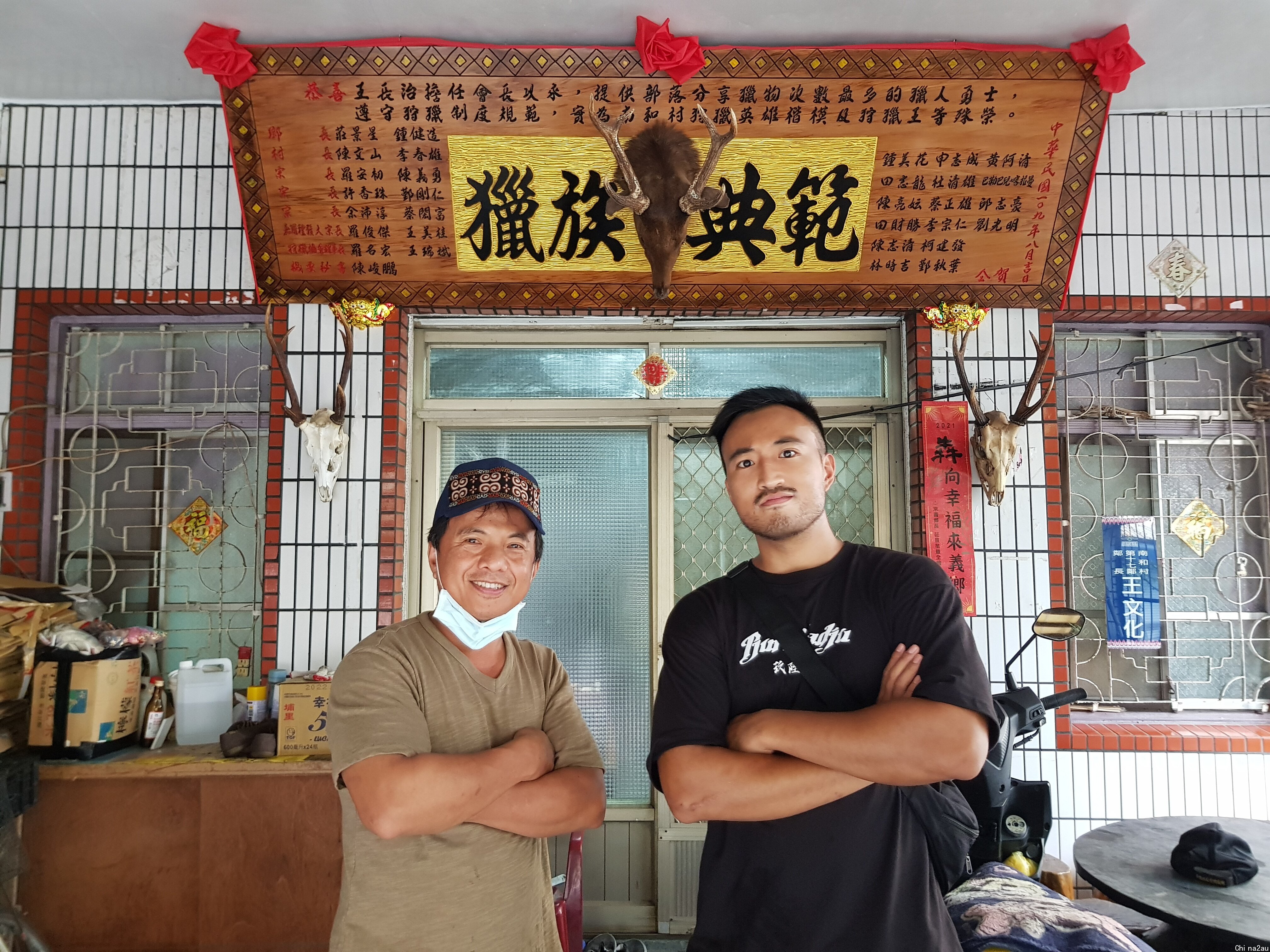 两名男子双手抱胸站在一块匾额下，匾额上用繁体中文书写“猎族典范”