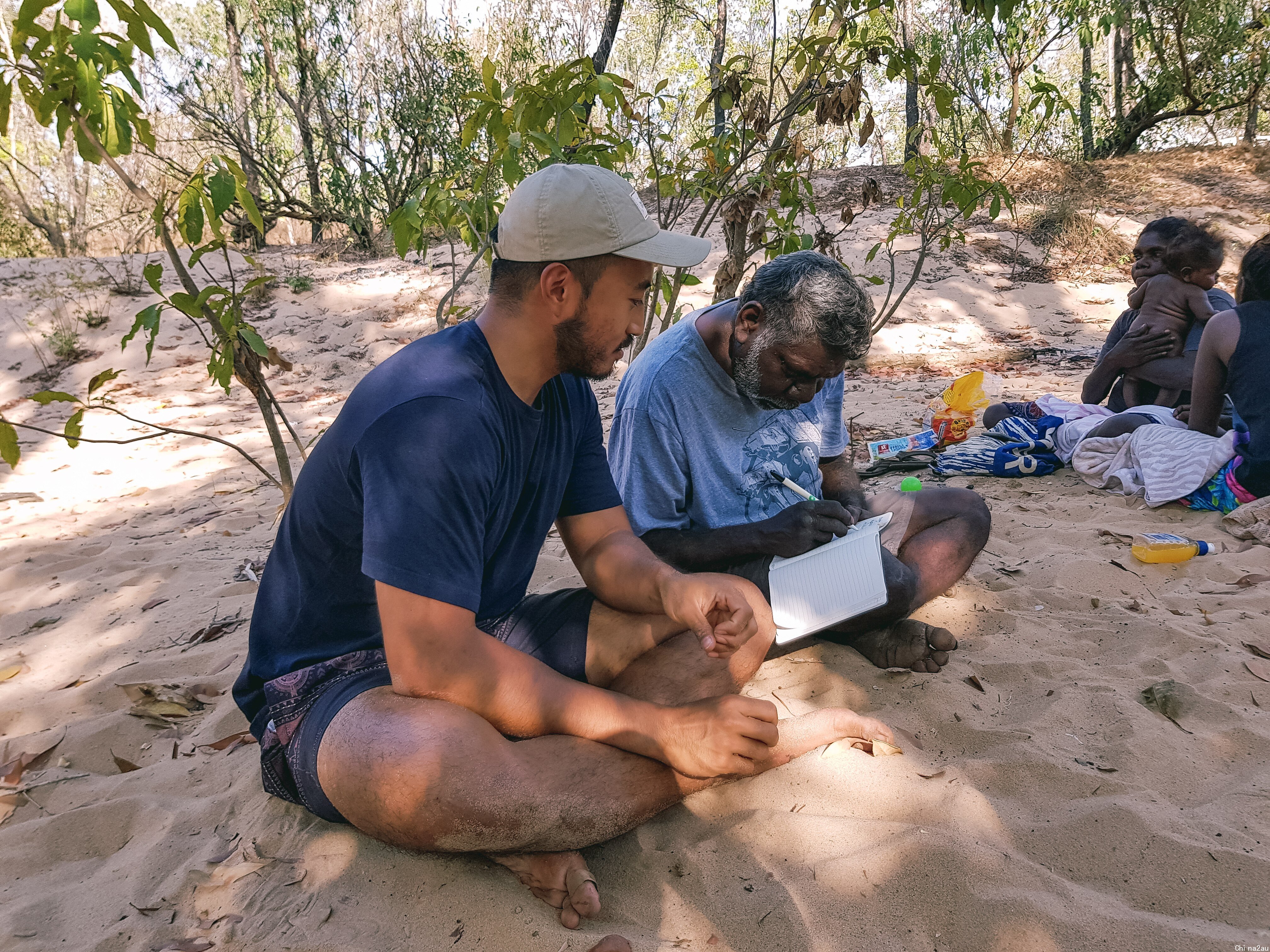 一名亚裔男子和一名原住民老人坐在一起，老人在笔记本上写字给亚裔男子看。