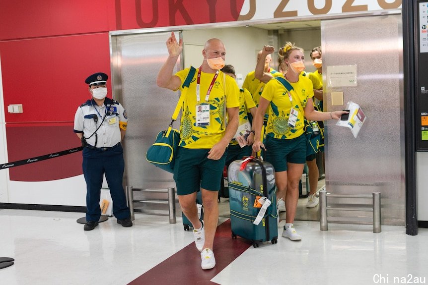 Australian athletes in green and gold tracksuits and face masks wave at an airport terminal 