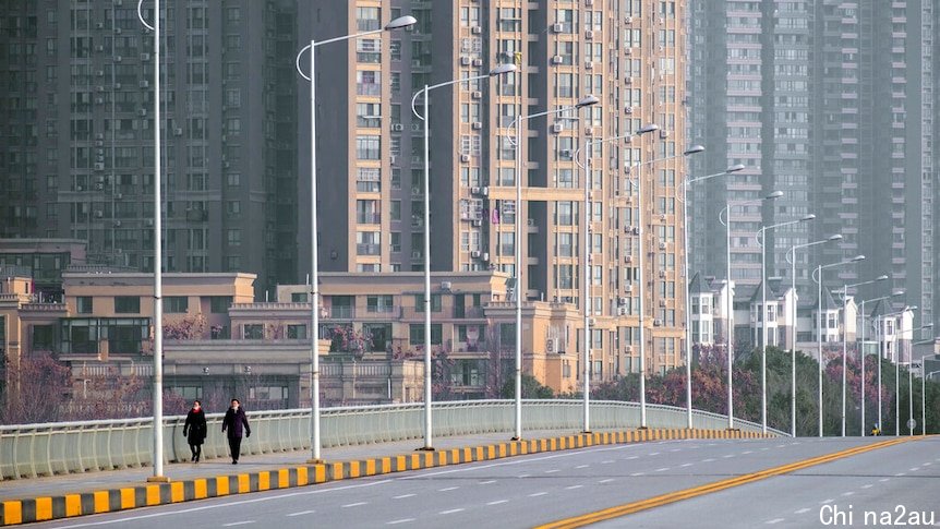 You view an empty six-lane bridge apart from two pedestrians with high rise buildings rising up behind them.