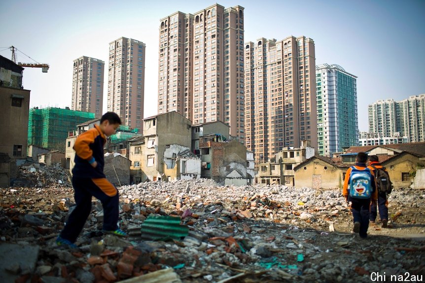 Three boys walk through rubble with a row of apartment towers along the skyline