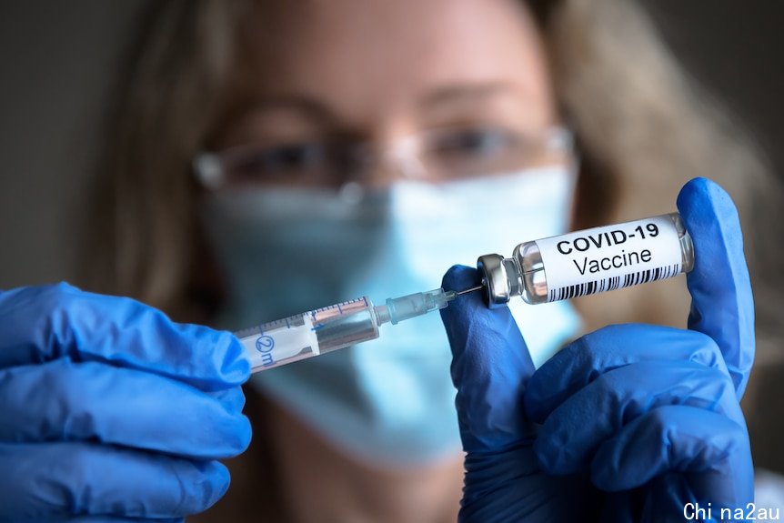 一位戴着外科口罩和蓝色手套的妇女举起一个COVID疫苗瓶和一个注射器。