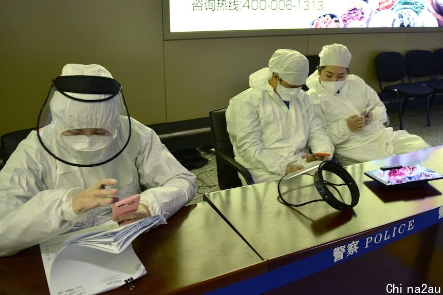 People wearing full PPE suits sit at a desk in an airport. A barrier in front of them says POLICE