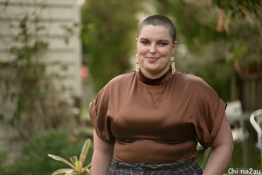 Noam O'Reilly wearing a brown blouse with shaved hair standing in her backyard