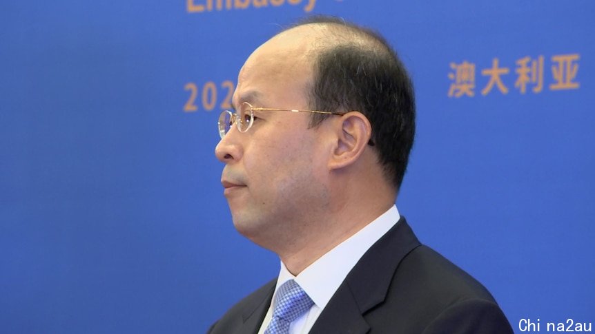 A profile shot of a middle-aged balding man wearing a suit and glasses, in front of a large backdrop.