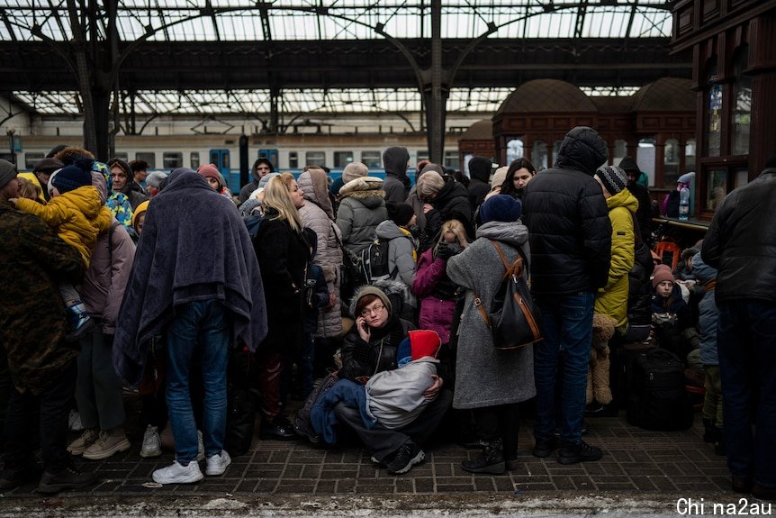 A train station platform, with people wearing blankets and winter clothes all huddled.