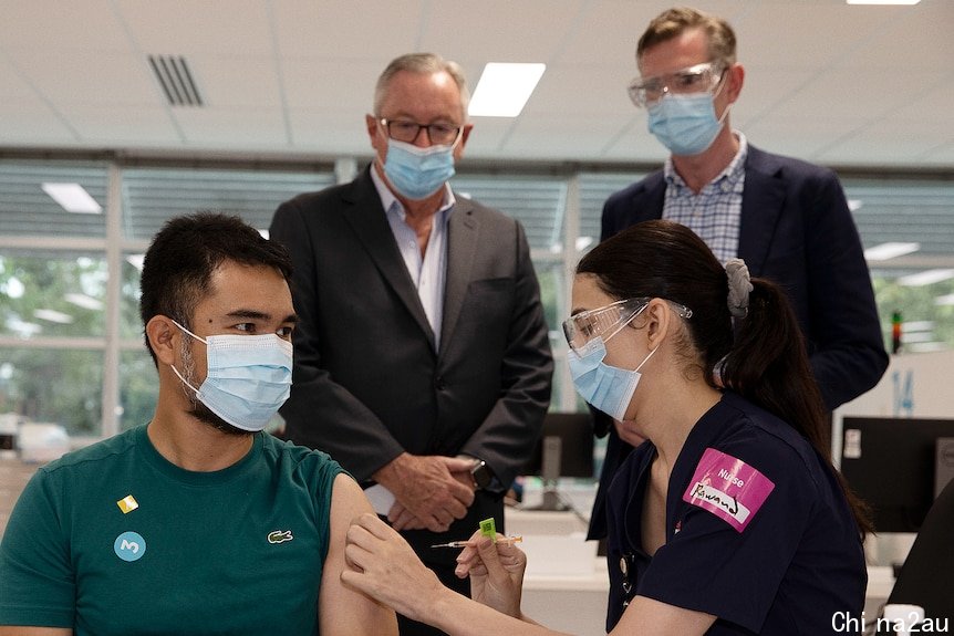 a man gets a vaccine while two other men watch on