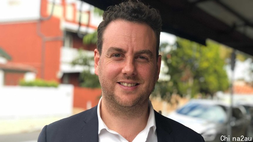 A man in his 30s wears a white shirt and dark suit and smiles at the camera.