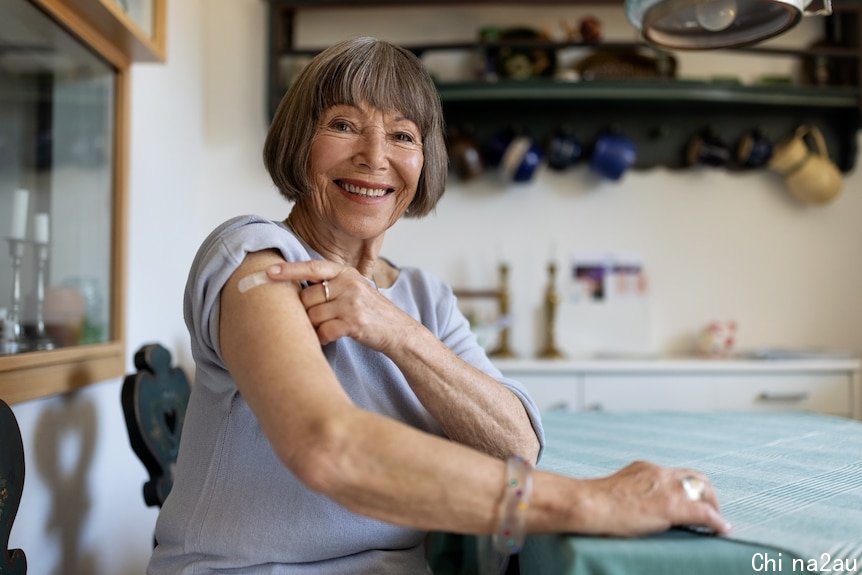 A senior white woman pointing to a bandaid on her upper arm and smiling