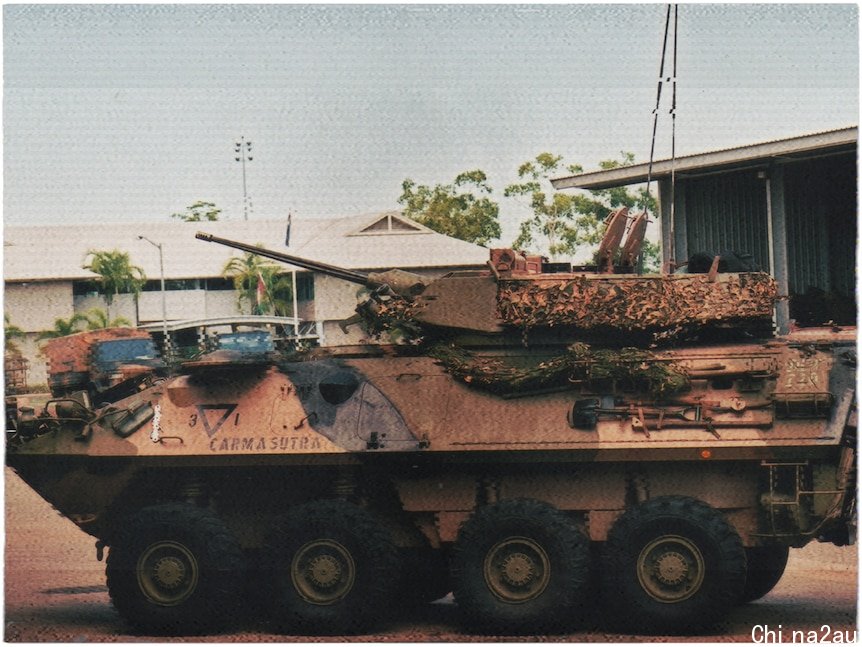 An eight-wheeled light armoured vehicle sits parked. Behind it are some low buildings and trees.