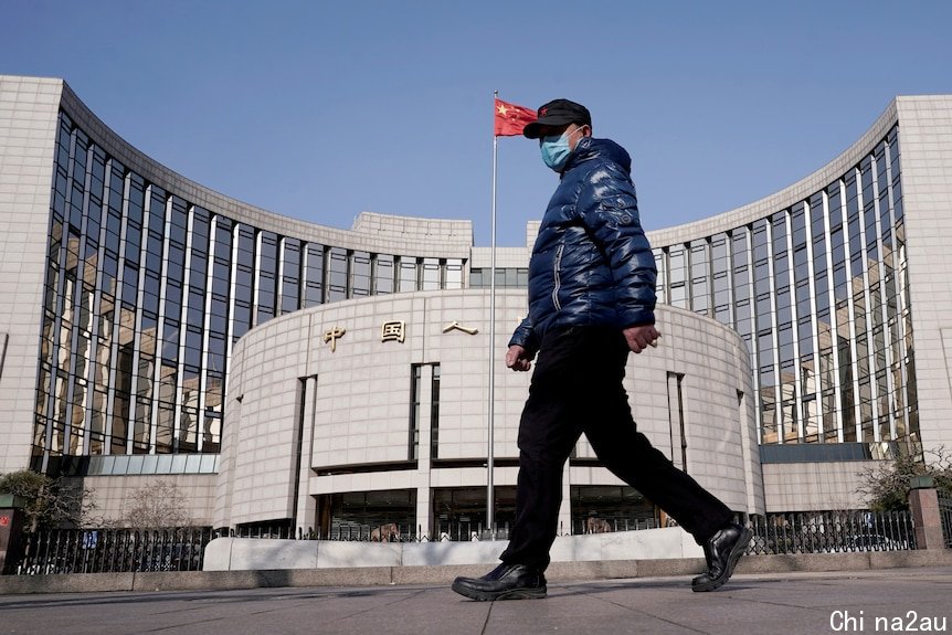 A man wearing a face mask and blue puffer jacket walks past a large building with a Chinese flag flying on a flagpole.