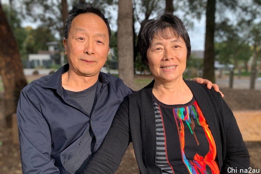 A Chinese couple smiling with the man's hand on the woman's shoulder and trees in the background.