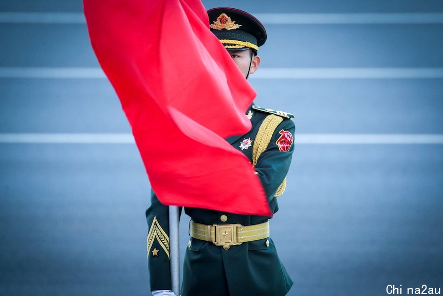A young man in Chinese military dress stands to attention with a red flag blowing across his face