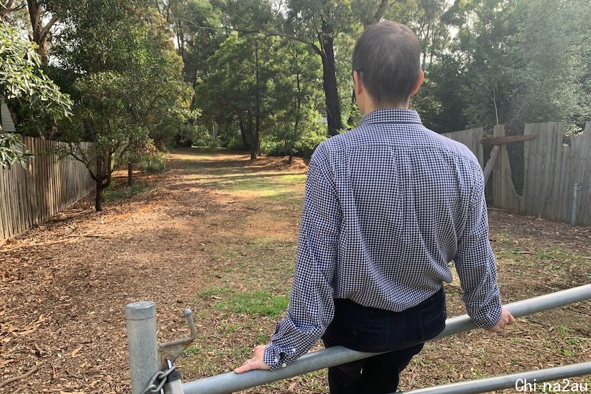 A man sitting on a metal barrier wearing a checked shirt looking towards trees.