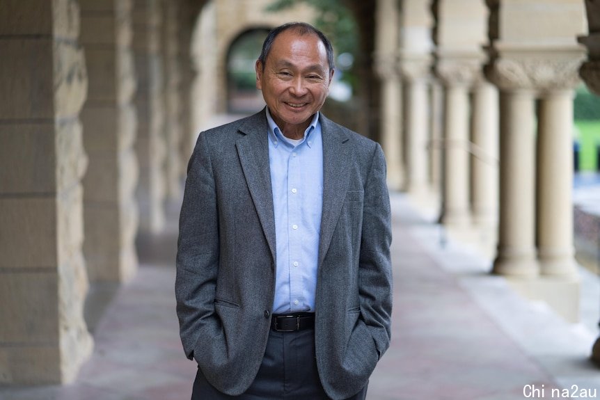 A man in a grey suit and blue shirt poses for a photo in a covered walkway flanked by sandstone columns