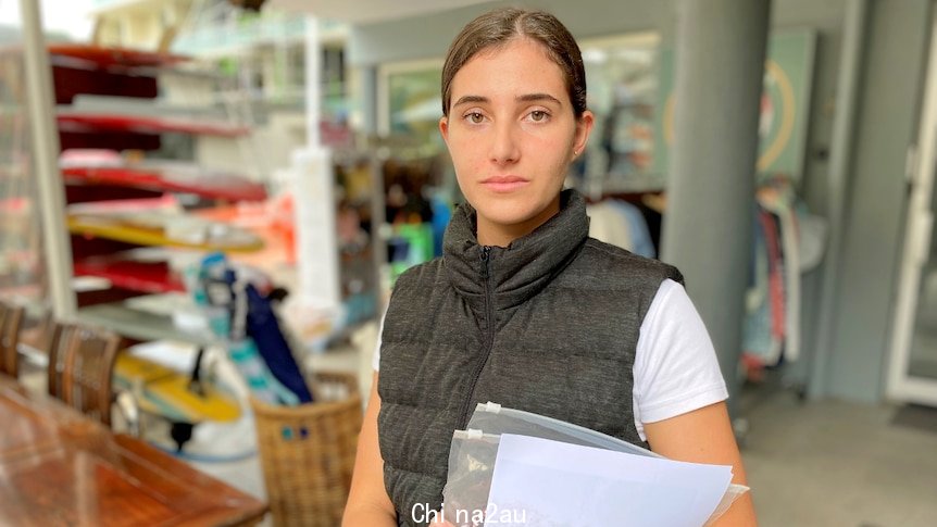 Woman stands holding papers, swimwear and not smiling