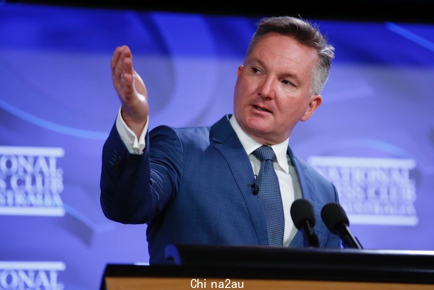 A man wearing a suit gestures as he stands in front of a lectern and a branded backdrop.