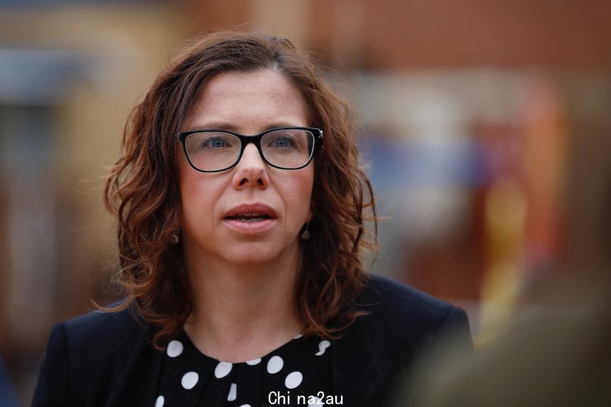 A woman with brown curly hair and glasses mid-sentence with everything else around her blurred out