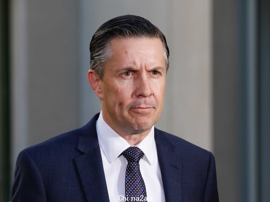 A man with slicked back brown hair wearing a suit and tie with a stern look on his face