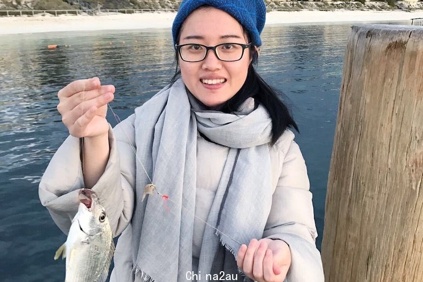 A woman holds a fish on a fishing line with water in the background.