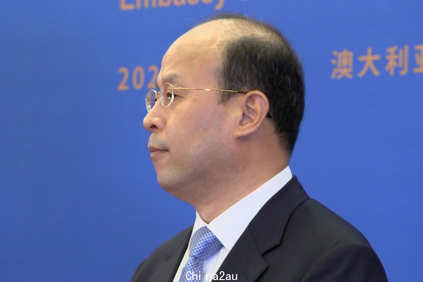 A profile shot of a middle-aged balding man wearing a suit and glasses, in front of a large backdrop.