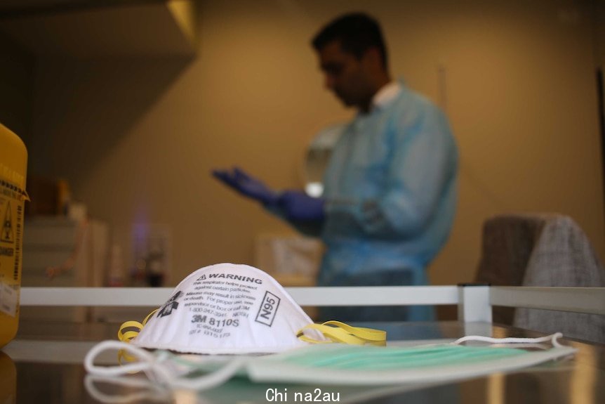 A doctor wears a blue gown and gloves in the background. A P2 mask sits in the foreground.