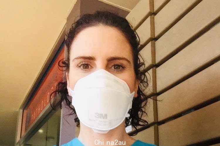 A close up profile image of Kerry with dark curly short hair, hazel eyes wearing white surgical mask infront of wall 