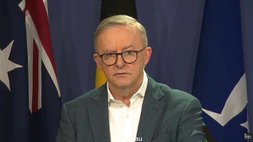 Anthony Albanese in a suit and glasses in front of australian, indigenous and torres strait island flags