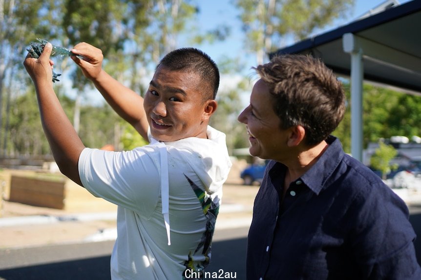 Young man wearing t-shirt standing outside holding a fresh tiger prawn in the air, a woman looks on smiling. 