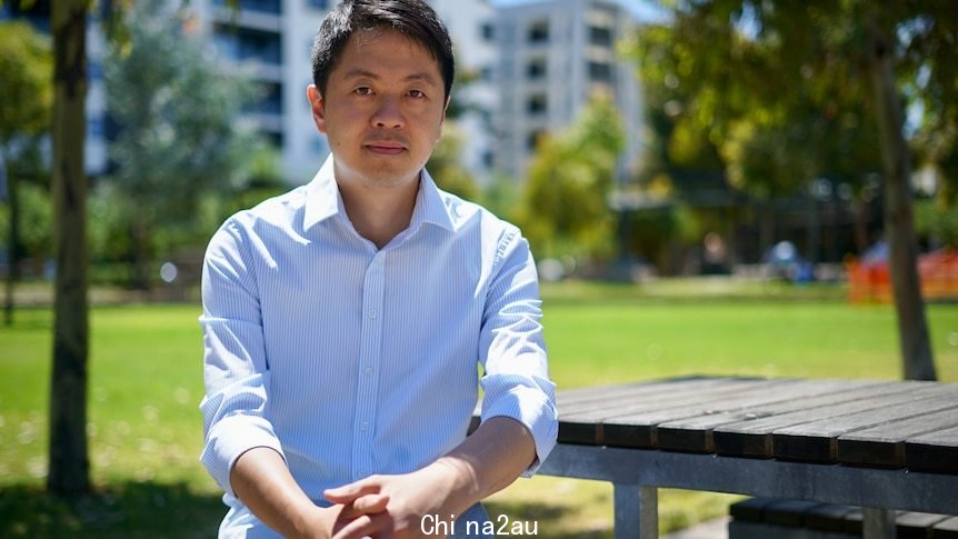 A man in a business shirt sits on a park bench surrounded by trees 