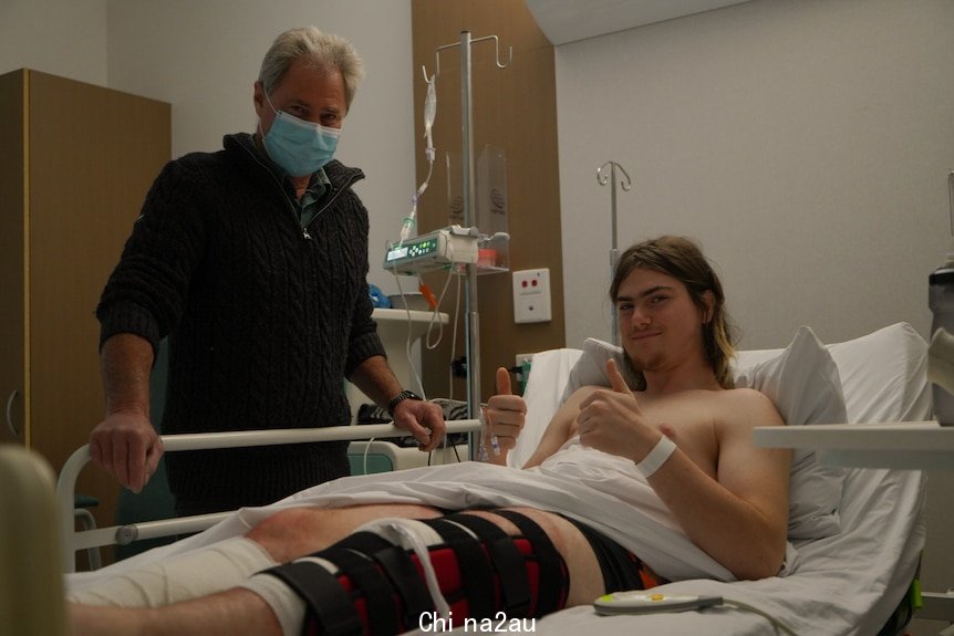 A teenager lying in a hospital bed with his father standing next to him.
