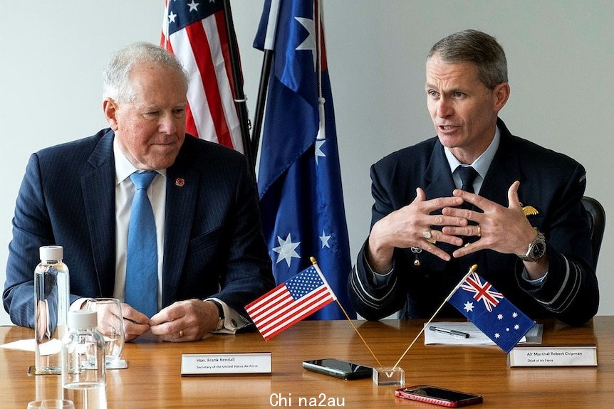 Two men in suits with military pins speak at a desk adorned with small United States and Australian flags