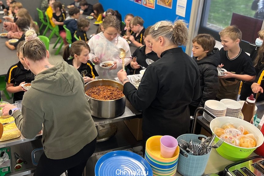 Lots of students sit at rows of tables to eat lunch while other children queue to get their taco bowls.