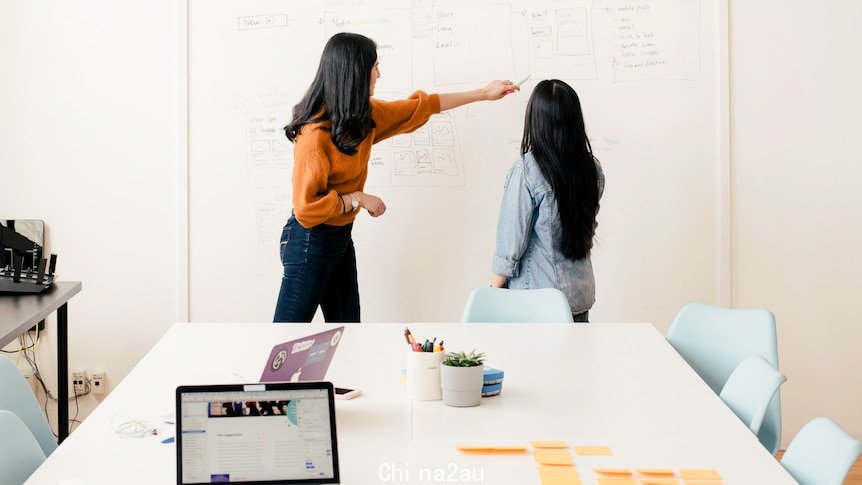 Two people stand in front of a whiteboard pointing at words.