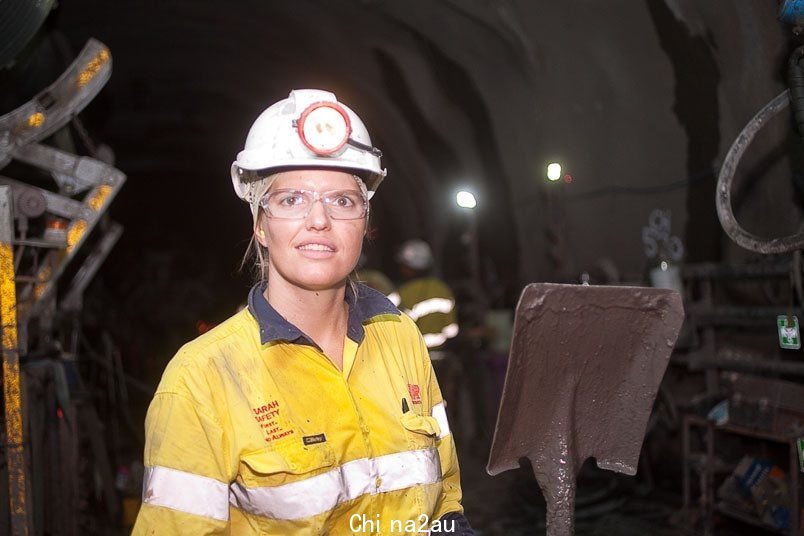 A woman with a hard hat on and high-vis shirt is in an underground environment smiling at the camera