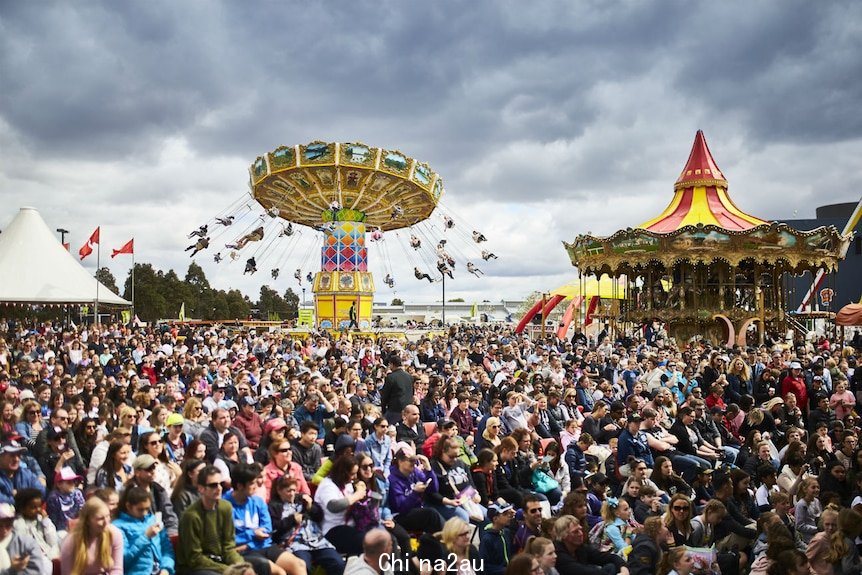 A large crowd gathers under a grey sky. There are rides and marquees in the background.