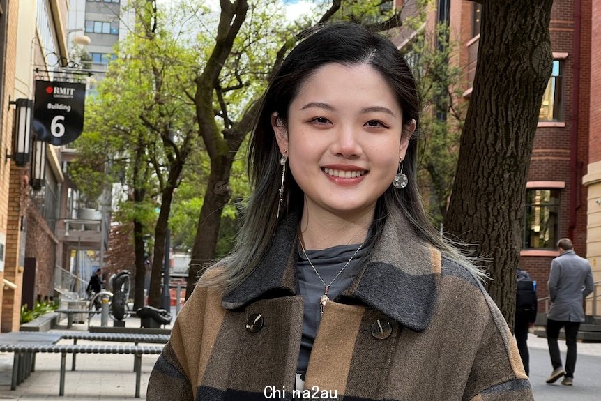 Jinru Sun wears a checked coat and smiles while standing under the trees outside RMIT