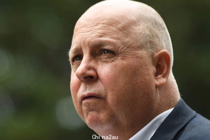 A man with a bald head wearing a suit jacket and shirt stands outside in profile.