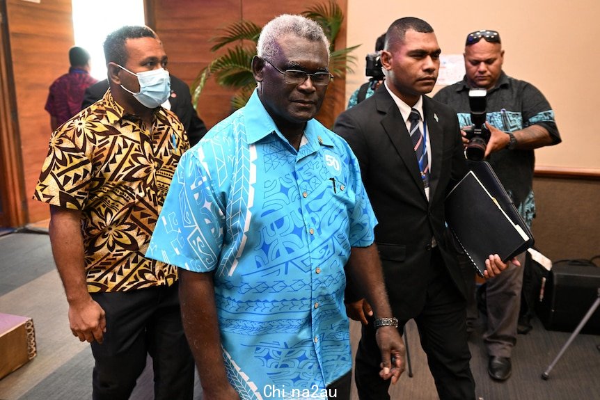 Man in blue shirt walks with two men beside as photographers take photos from behind.