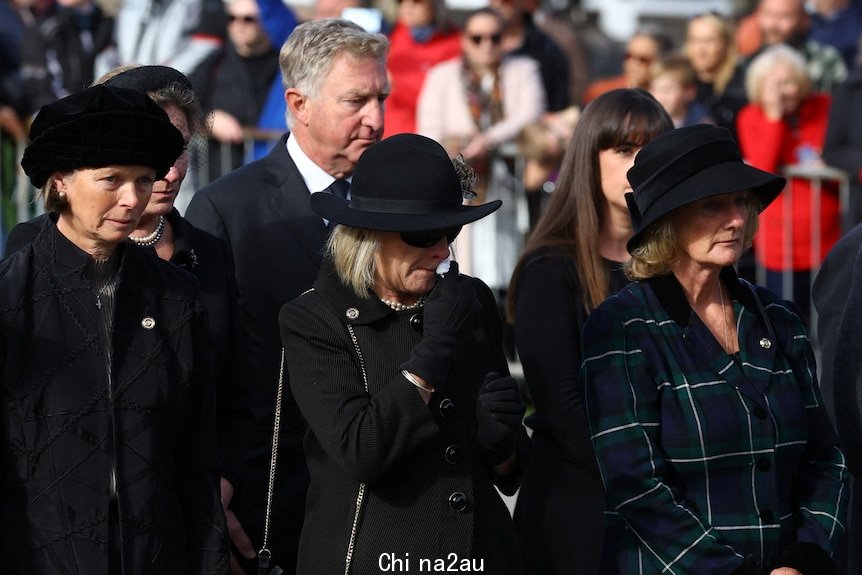 Three women in black clothing appear upset as they watch the hearse pass.