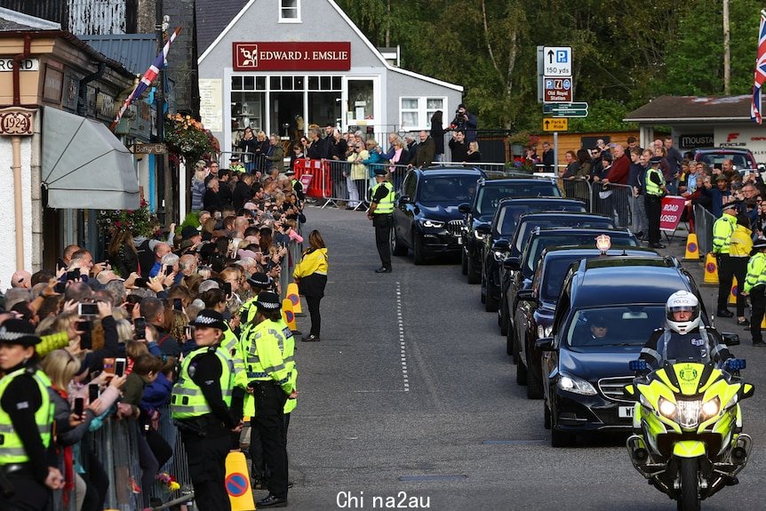 Crowds of people behind barricades and police in hi-vis as the funeral procession drives through street