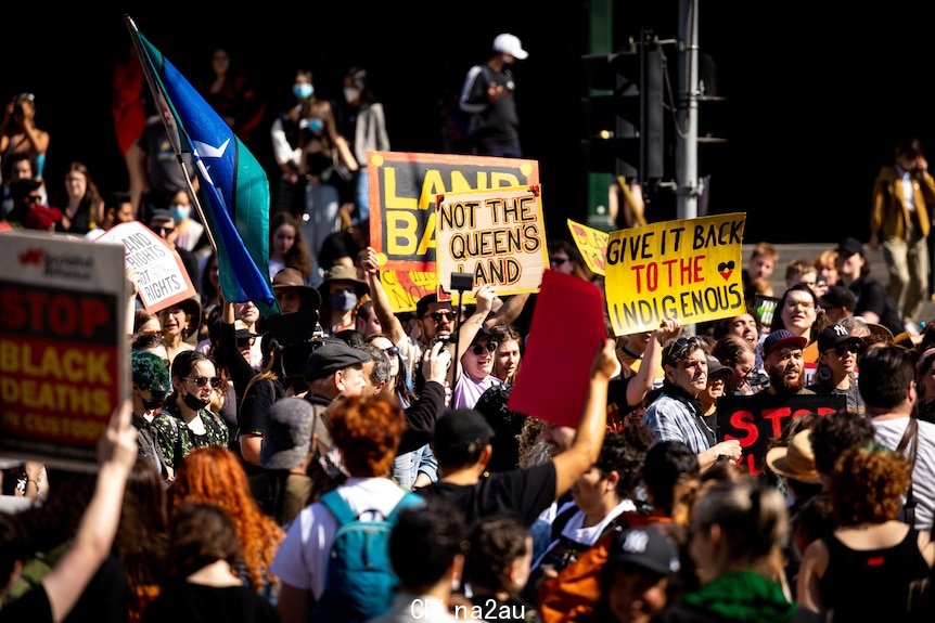 A crowd of people protesting, with some people holding signs saying 'not the Queen's land' and holding flags.