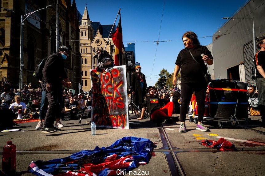 An Australian flag doused in fake blood at an anti-monarchy protest, with a sign saying 'stop black deaths in custody' behind.