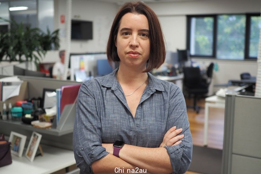 Woman with dark mid length hair, wearing grey shirt, with sleeves rolled up, unsmiling, arms crossed and standing in open office