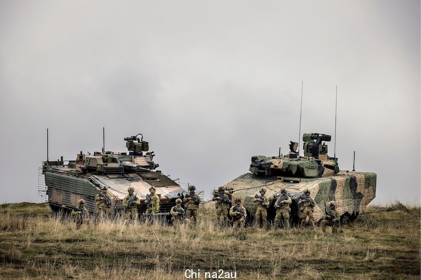 A group of 12 soldiers in combat gear pose with their rifles in front of two large, heavy, tank-like combat vehicles in a field.