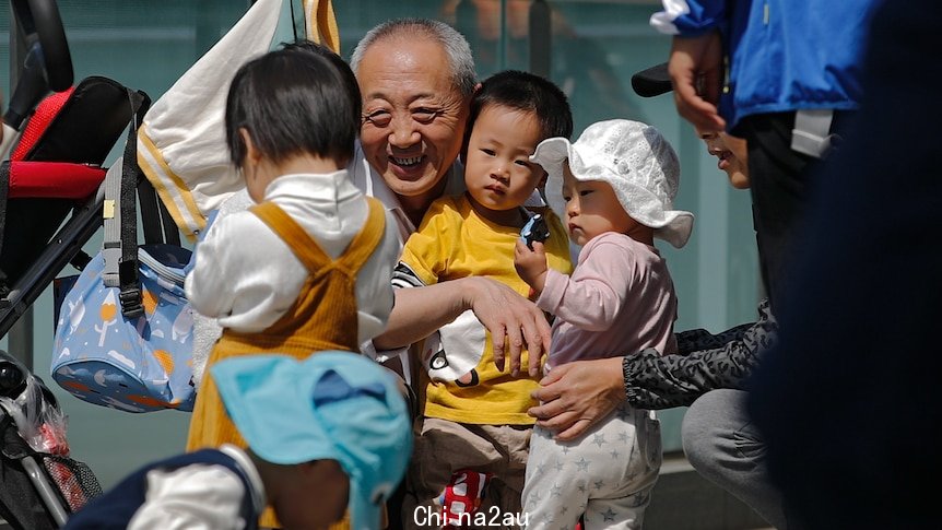 An elderly man smiles widely as he squats to hug and play with three small children in front of an office building.